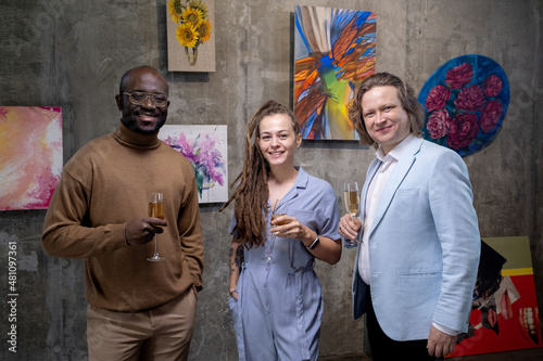 Three happy intercultural guests of art gallery toasting with champagne while standing against collection of paintings on wall