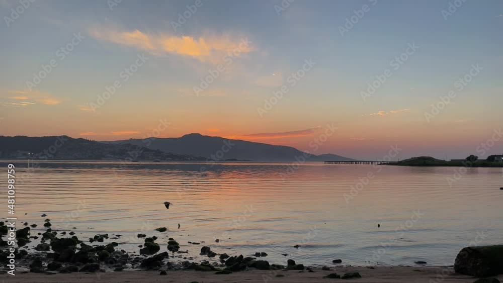 sunrise by the beach, sea with calm waters, sky in different colors, mountains in the background