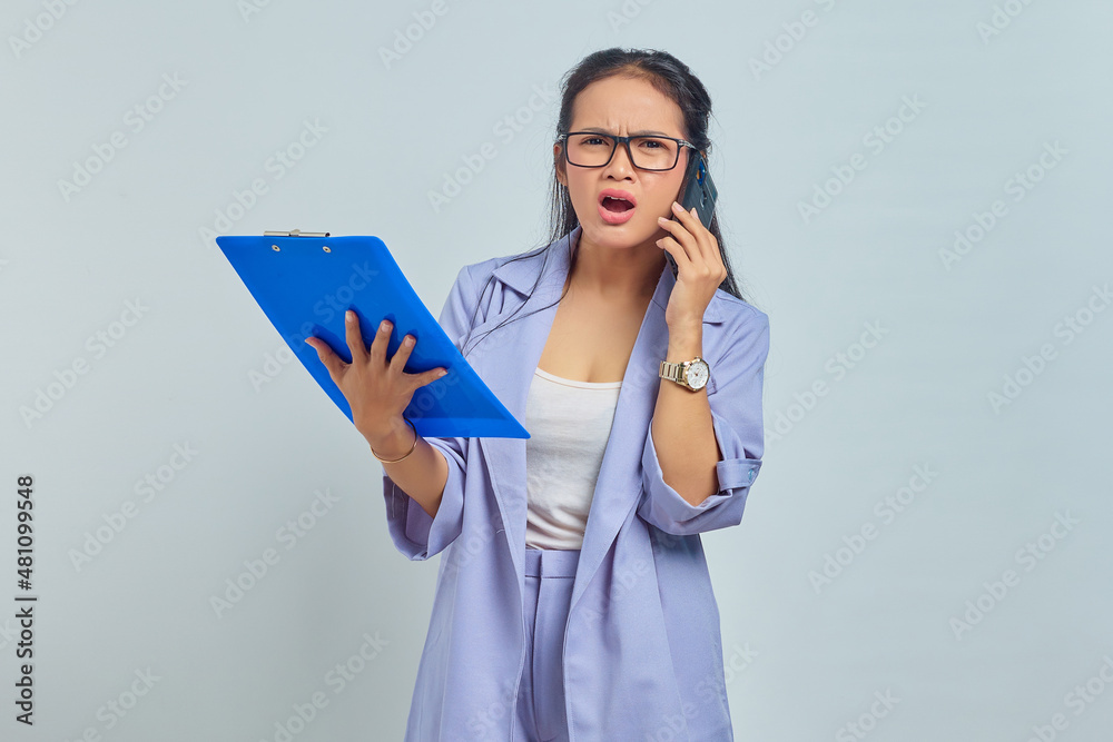Portrait of surprised young Asian woman talking on mobile and looking at task list in documents folder isolated on purple background