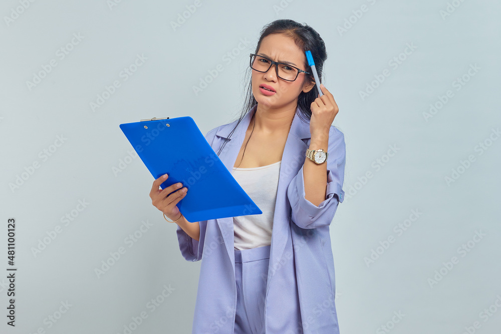 Portrait of young Asian business woman scratching head with pen, thinking of ideas and holding clipboard isolated on purple background