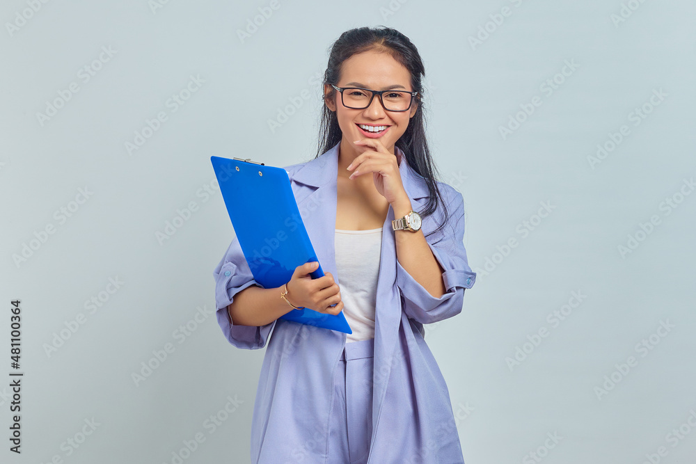 Portrait of cheerful young Asian business woman holding chin while holding document folder isolated on purple background Portrait of cheerful young Asian business woman holding chin while holding document folder isolated on purple background