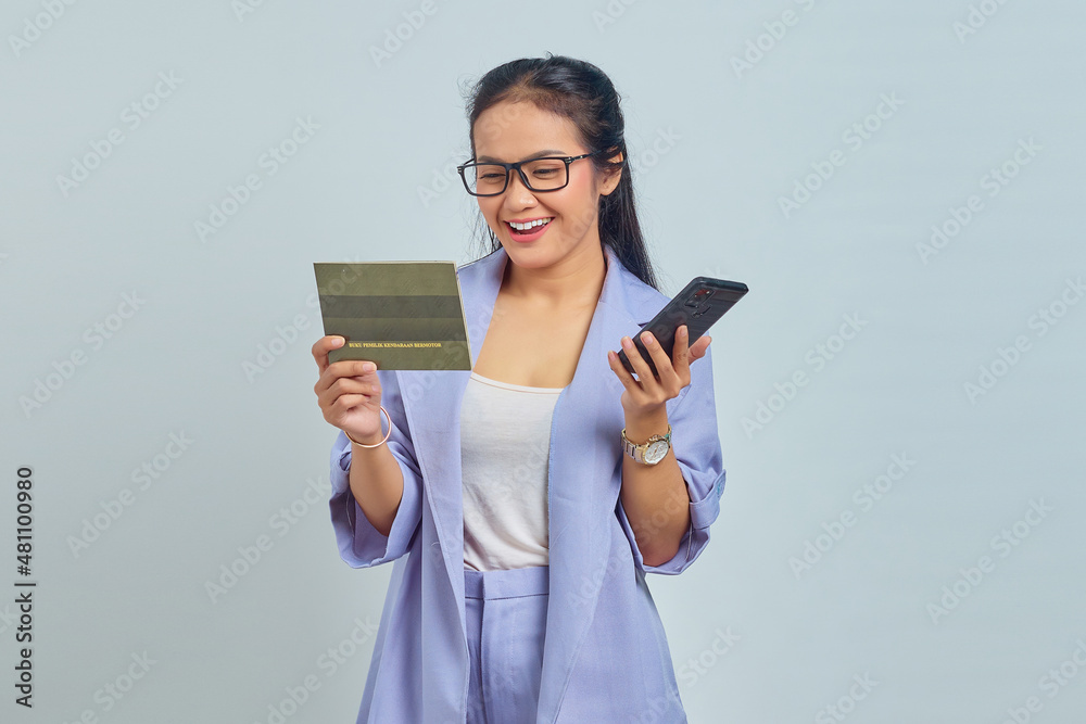 Portrait of cheerful young Asian woman holding mobile phone and looking at vehicle book isolated on white background Portrait of cheerful young Asian woman holding mobile phone and looking at vehicle book isolated on white background
