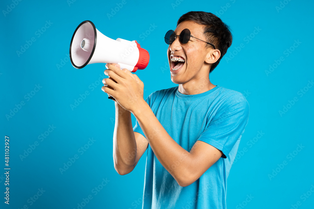 Handsome young man over isolated blue background shouting through a megaphone Handsome young man over isolated blue background shouting through a megaphone