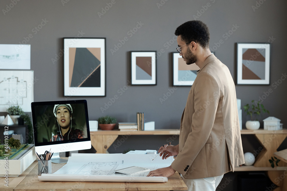 Young biracial engineer in formalwear looking at female builder on computer screen during online discussion of working points Young biracial engineer in formalwear looking at female builder on computer screen during online discussion of working points