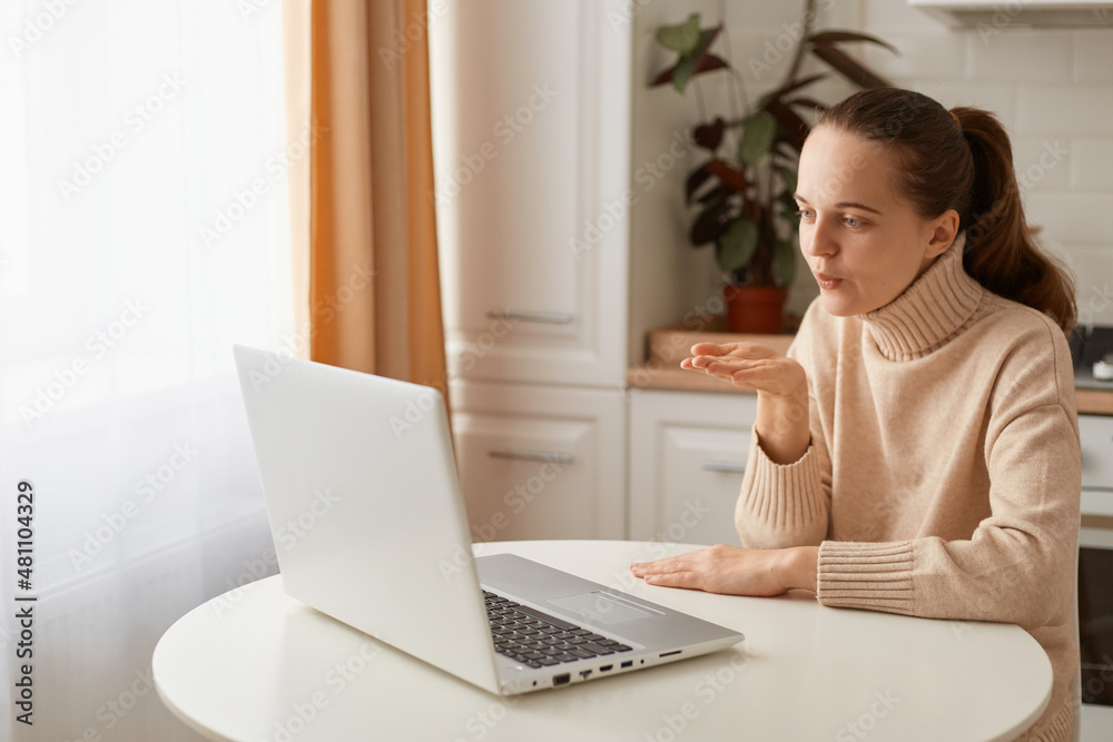 Portrait of beautiful young adult woman with ponytail hairstyle wearing casual style beige sweater sitting at table in kitchen and having video call via notebook, sending air kiss to laptop display. Portrait of beautiful young adult woman with ponytail hairstyle wearing casual style beige sweater sitting at table in kitchen and having video call via notebook, sending air kiss to laptop display.