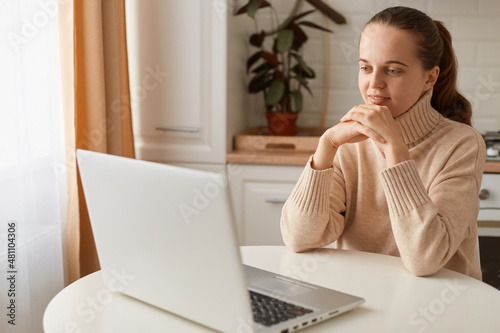 Portrait of positive woman with ponytail hairstyle wearing casual style beige sweater sitting at table in kitchen and looking at laptop screen, holding hands under chin, watching movie or webinar.