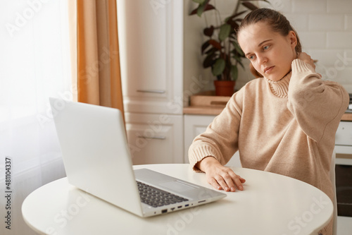 Portrait of tired woman with ponytail hairstyle wearing casual style beige sweater sitting at table in kitchen and working online on computer, feels pain in neck after long hour work, looks exhausted.