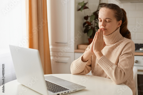 Indoor shot of relaxed woman with ponytail wearing casual style beige sweater sitting at table in kitchen in front of computer, keeping hands in praying gesture, keeps eyes closed, trying to rest.