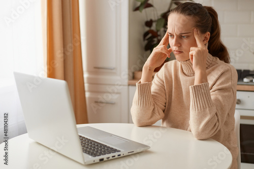 Indoor shot of pensive woman wearing beige casual style sweater with ponytail hairstyle sitting at table in front of notebook, thinking over new project, keeping fingers on temples.