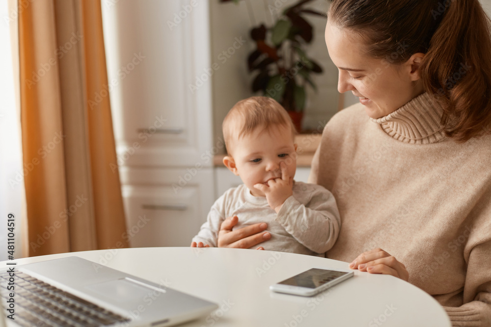 Indoor shot of beautiful Caucasian woman wearing beige casual style sweater with ponytail hairstyle sitting at table with her baby daughter and looking at baby with charming smile and gentle. Indoor shot of beautiful Caucasian woman wearing beige casual style sweater with ponytail hairstyle sitting at table with her baby daughter and looking at baby with charming smile and gentle.