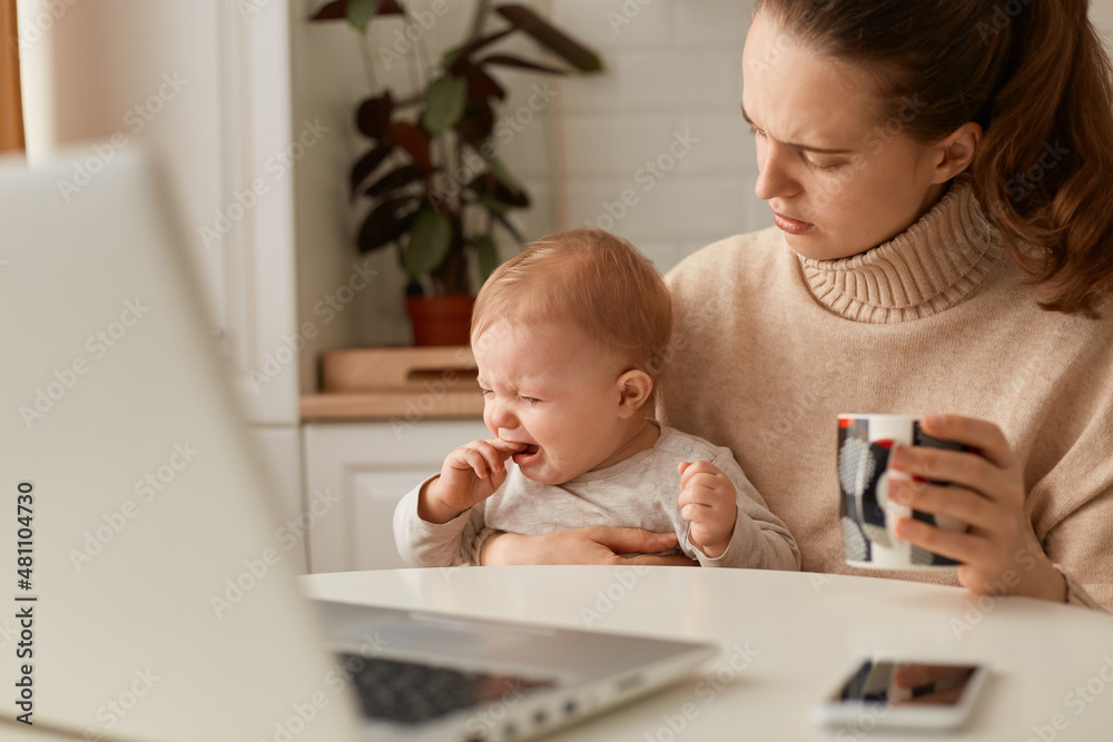 Portrait of sad woman wearing beige casual style sweater with ponytail hairstyle sitting at table with her crying infant daughter, holding cup of coffee or tea, trying to calm down baby. Portrait of sad woman wearing beige casual style sweater with ponytail hairstyle sitting at table with her crying infant daughter, holding cup of coffee or tea, trying to calm down baby.