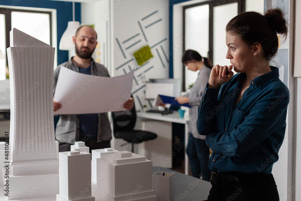 Architect looking into the distance gazing at skyscraper scale model with team colleague engineer holding construction plans. Team of architects brainstorming architectural ideeas. Architect looking into the distance gazing at skyscraper scale model with team colleague engineer holding construction plans. Team of architects brainstorming architectural ideeas.