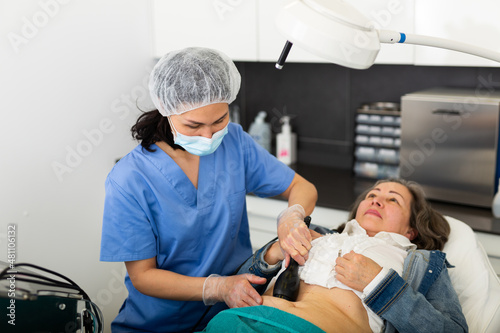 Asian woman professional cosmetologist performing abdominal skin rf lifting procedure on modern equipment for aged female patient in aesthetic medicine office