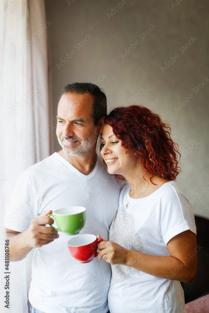 Cute middle aged european couple tender hugs and morning coffee together. Man and woman together at the window in the bedroom, lifestyle in a real interior. Support and emotions Cute middle aged european couple tender hugs and morning coffee together. Man and woman together at the window in the bedroom, lifestyle in a real interior. Support and emotions