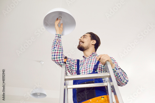 Electrician screwing in light bulb at home. Happy man in work uniform standing on ladder in residential building, office, school or hospital and changing LED lightbulb in modern white lamp on ceiling