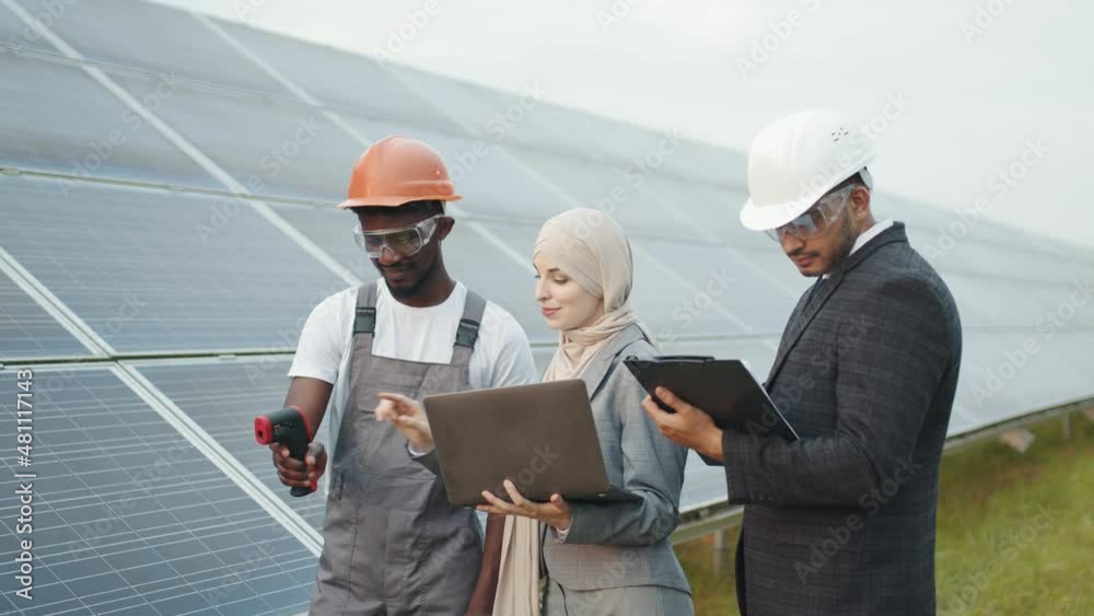 Multiracial people controlling temperature of solar panels. Muslim woman with laptop and indian man with clipboard standing on solar farm with african american technician. Industrial worker. Multiracial people controlling temperature of solar panels. Muslim woman with laptop and indian man with clipboard standing on solar farm with african american technician. Industrial worker.