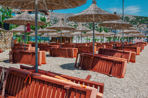 View of empty beach – wooden sunbeds and shade umbrellas near sea water, city buildings and mountains on the horizon. Landscape of abandoned summer resort.