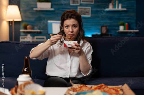 Tired woman holding chopsticks and chinese takeaway noodle box sitting on couch after long day at the office. Person looking sad eating delivery asian fast food watching evening news.