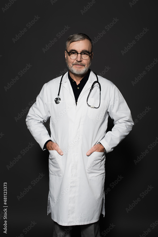 smiling senior doctor looking at camera while standing with hands in pockets on dark grey. smiling senior doctor looking at camera while standing with hands in pockets on dark grey.