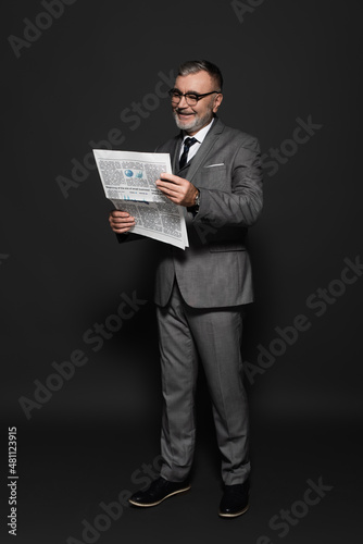 full length of senior man in suit and eyeglasses reading newspaper on dark grey.