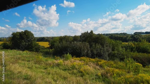 View to the countryside yellow fields from open window of quickly driving train. Rural summer meadow landscape at sunny summer day on a blue cloudy sky background.