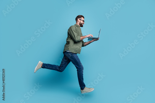 Profile photo of nice freelancer guy jump hold netbook typing email wear specs green shirt isolated blue color background