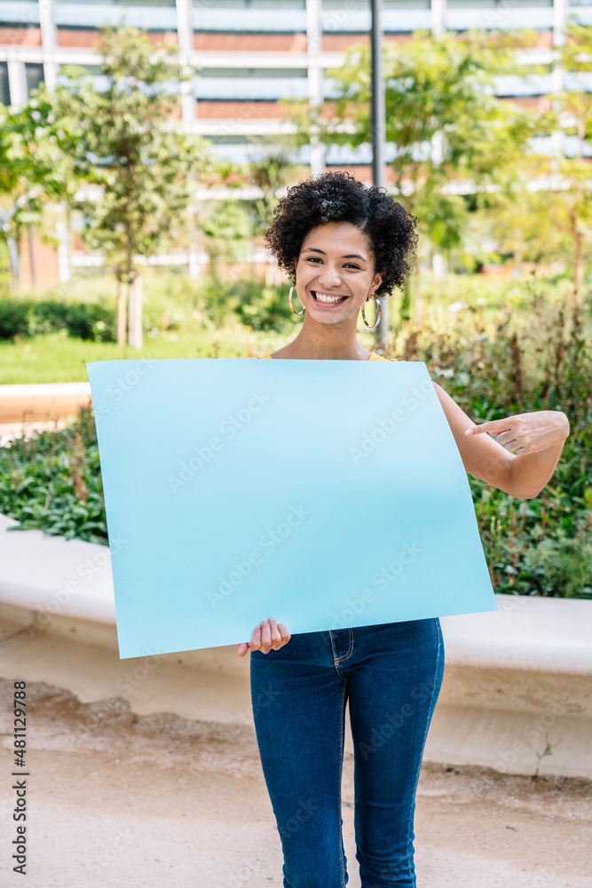 Positive young ethnic woman pointing at empty billboard and smiling in park Positive young ethnic woman pointing at empty billboard and smiling in park