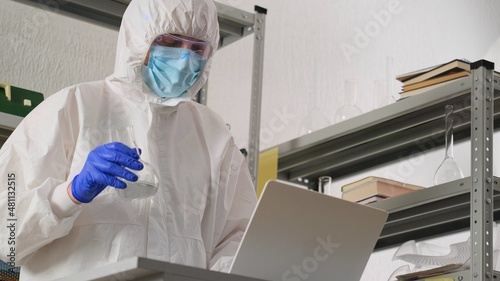 Man scientist or medical working  in protective uniform  looking on flask with liquid in science lab and recording results to laptop