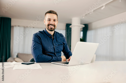 A happy businessman typing on the laptop, working from home and smiling at the camera.