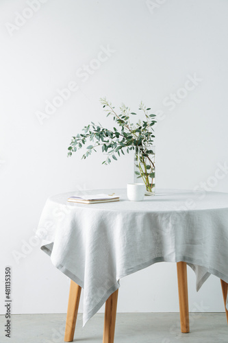 Side view round table with a rough white tablecloth and branch in a jar. Cropped, over white wall. Indoors. Notepad and cup on a surface.
