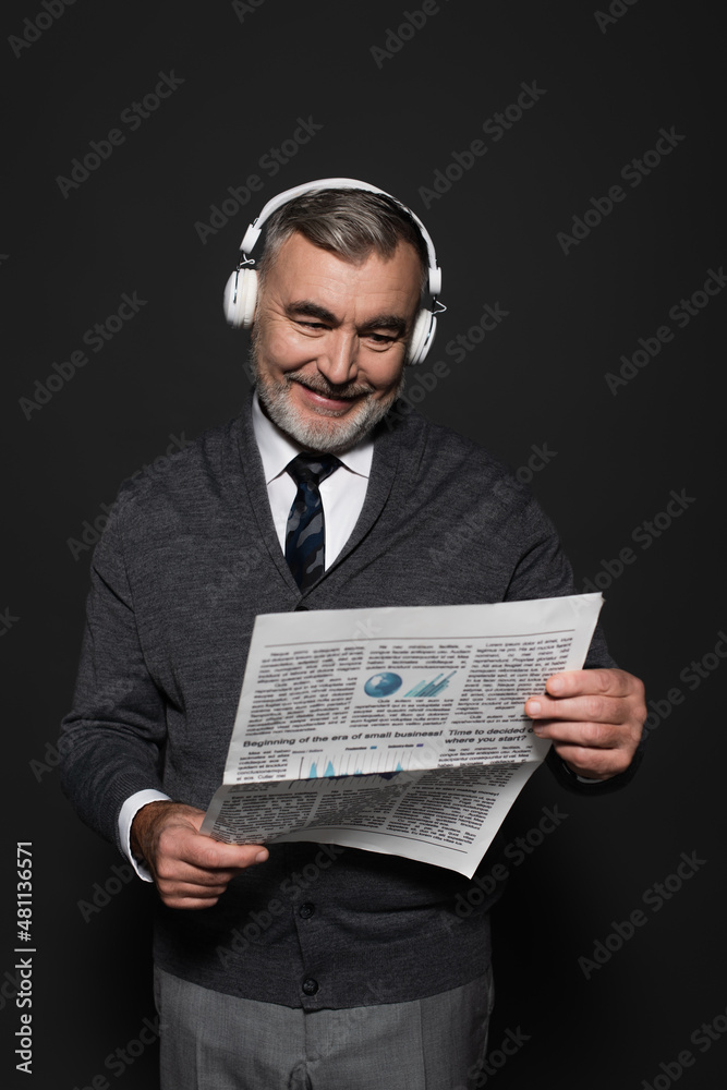 happy senor man in jumper, tie and headphones reading newspaper on dark grey. happy senor man in jumper, tie and headphones reading newspaper on dark grey.