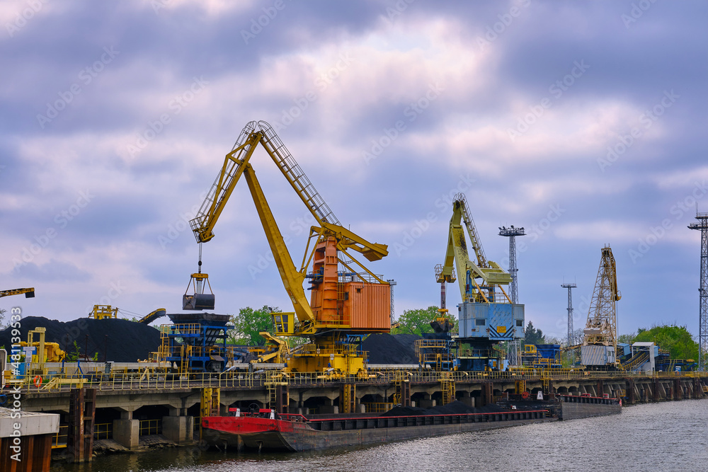 River port cranes with clamshell or griper loading coal to river drag boats or barges moored by pier on cloudy day. Energy, minerals, environment River port cranes with clamshell or griper loading coal to river drag boats or barges moored by pier on cloudy day. Energy, minerals, environment
