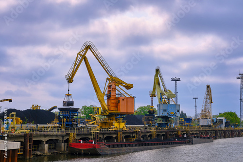 River port cranes with clamshell or griper loading coal to river drag boats or barges moored by pier on cloudy day. Energy, minerals, environment 