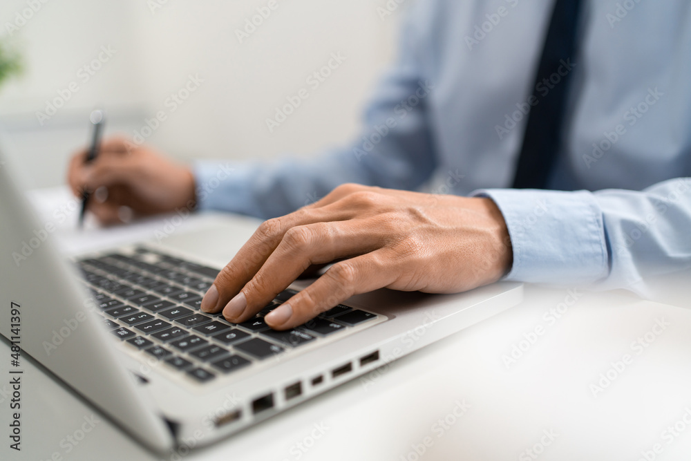 Working Man Conept The male officer sitting in the office, laying his left hand on the laptop, and writing something on the paper with his right hand Working Man Conept The male officer sitting in the office, laying his left hand on the laptop, and writing something on the paper with his right hand