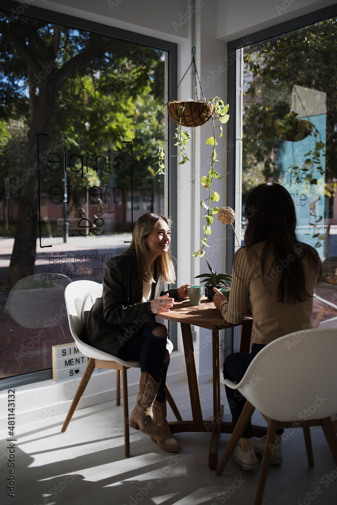 Two women chat and laugh while having a cup of tea. They are sitting in a corner of a very bright cafeteria with large windows. Translation Two women chat and laugh while having a cup of tea. They are sitting in a corner of a very bright cafeteria with large windows. Translation