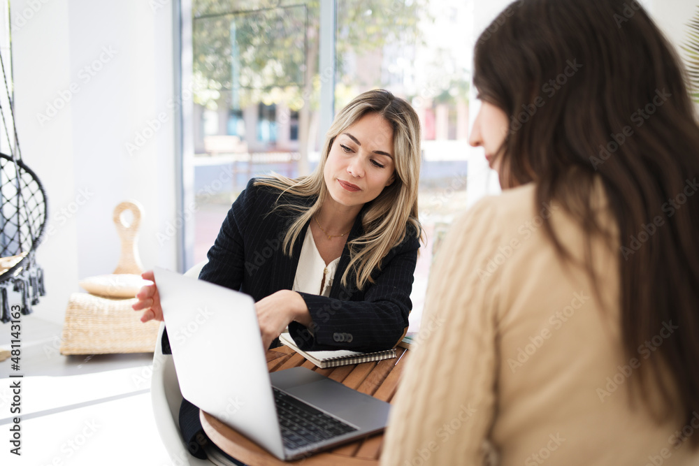 Two women have a business meeting inside a coworking space. One of them shows her something on the laptop. Two women have a business meeting inside a coworking space. One of them shows her something on the laptop.