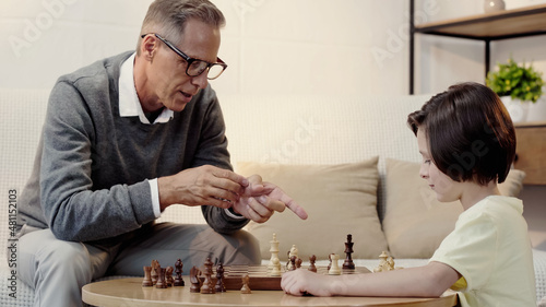 grandfather in glasses pointing at figure while playing chess with boy in living room