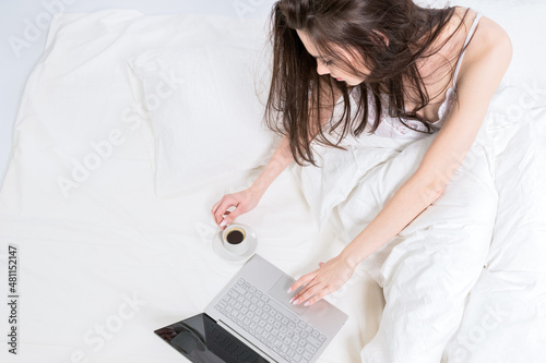 A cute brunette is sitting on a white bed next to a laptop and a cup of coffee. Good morning. Work from home