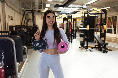 Portrait of smiling trendy 20 years old gym owner woman, holding mats and showing open sign. Opening fitness centar after covid-19 pandemic. People and healthcare concept. Copy space. photo