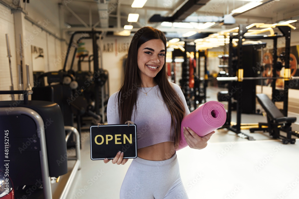 Obraz premium A beautiful young girl, looking at the camera with a smile on her face, holds a sign that the gym has reopened after a long time. A young woman holding a yoga mat indicates that the gym has reopened.