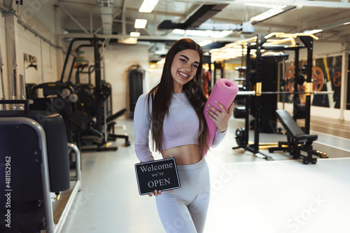 Cropped shot of a gym instructor holding up a "open sign". Attractive young woman opening up her fitness center. Portrait of a sporty young woman holding an exercise mat and "open" sign in a gym. © Jelena Stanojkovic