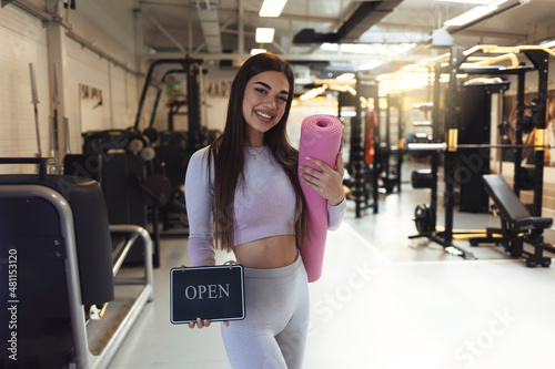 Portrait of smiling trendy 20 years old gym owner woman, holding mats and showing open sign. Opening fitness centar after covid-19 pandemic. People and healthcare concept. Copy space. photo