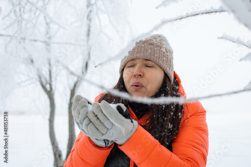 Woman of middle age behind some twigs covered with snow blowing snow off gloves on walk in snowy forest with tree in background. Magic winter time full of white color. 