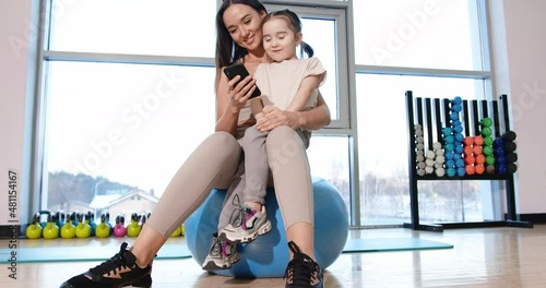 An attractive mother with her daughter are sitting on a gymnastic ball and looking at a mobile phone. Sporty young family. BMPCC 6k.