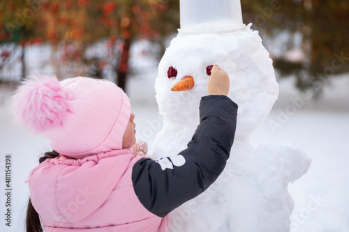 Young girl alone making face of snowman with improvised means in daytime while having walk in park with trees and bright sun light in background. Parents spending time with children. photo