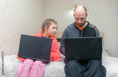 Dad and daughter are sitting on the bed with laptops on their laps. A man and a child work on computers at home. Daughter peeps into dad's screen photo