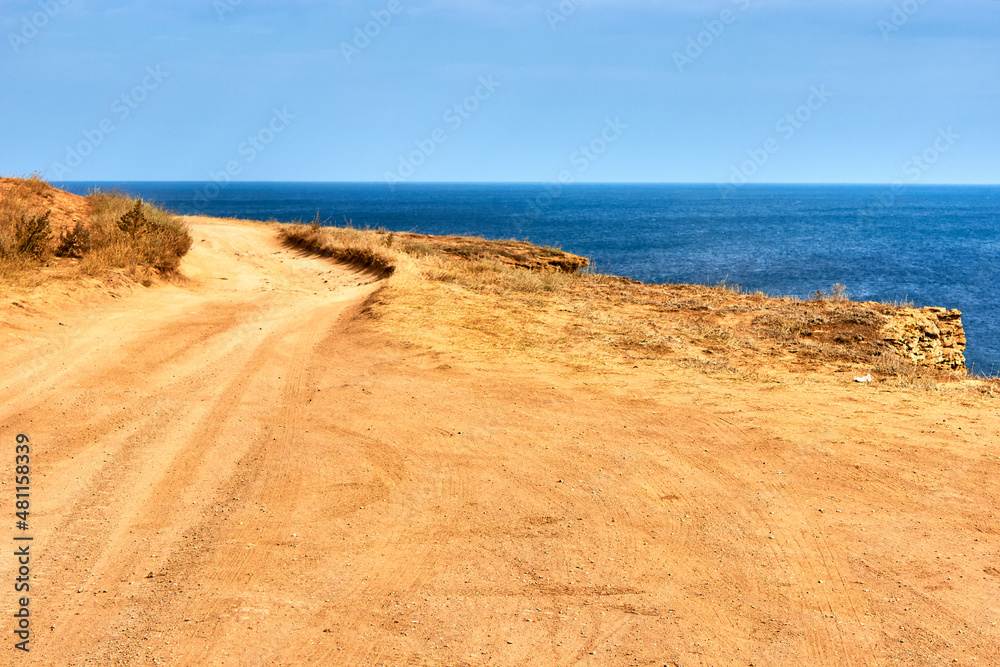 A sandy country road along the edge of the cliff. Orange land, a cape by the sea. Seascape, horizon, calm blue sea, clear sky.