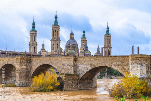 Ancient Stone Bridge of Lions on the backdrop of the towers of Cathedral-Basilica of Our Lady of the Pillar in Saragossa, Spain. Medieval cityscape with Ebro river with yellow water on an autumn day