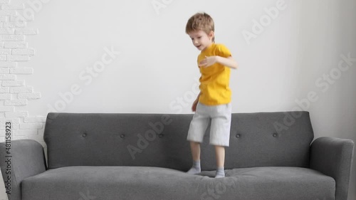 Lovely happy little boy having fun alone while jumping on the soft gray couch in contemporary apartment expressing happiness and positive emotion, slow motion