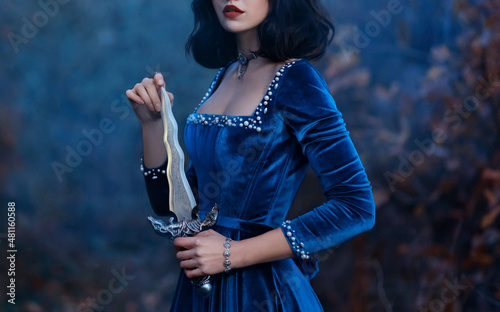 face cropped, red lips close-up. Fantasy medieval woman warrior queen holding dagger, knife in hands. velvet vintage blue dress. Girl princess vampire, brunette short hair. Nature forest dusk night photo
